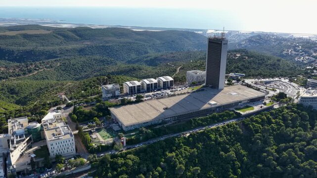Drone flight capturing an elevated orbiting perspective of the modern Eshkol Tower and institutional buildings situated on the green forested ridge of Mount Carmel.