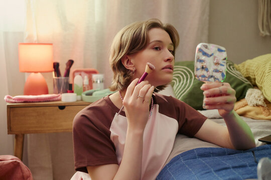 Young girl applying makeup while checking handheld mirror in bedroom, expressing self care and identity exploration. Useful for beauty, youth lifestyle, skincare, wellness marketing