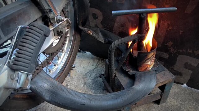 A rubber motorcycle inner tube is clamped onto a metal vulcanizing press next to a burning container with open flames, positioned beside a vehicle wheel on a workshop floor.