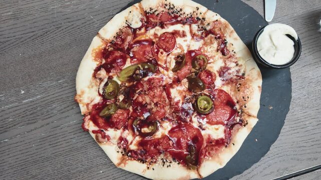 Delicious pepperoni pizza topped with jalape&ntilde;os and cheese on a black slate plate, served with creamy dip on a rustic wooden table. Overhead, static close-up shot, soft natural lighting.