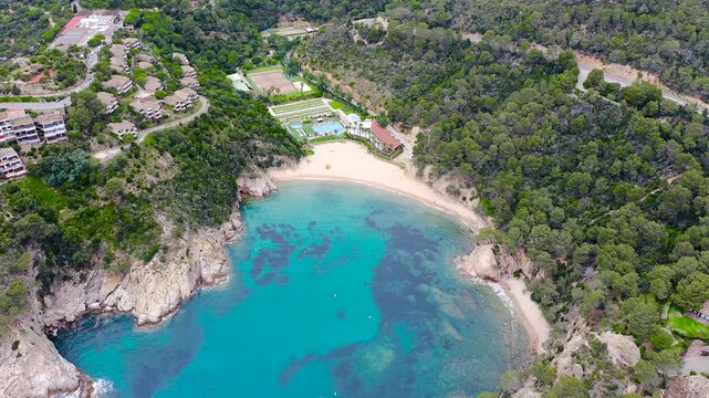 Aerial view of Cala Giverola, a secluded Mediterranean cove with crystal-clear turquoise waters, anchored boats, rugged cliffs and pine forests along the Costa Brava, Girona. 