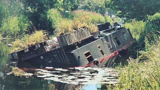 A static B-roll shot showing a large, dark-colored traditional narrowboat houseboat that has partially sunk and capsized onto its side in the Grand Canal of Dublin, Ireland.