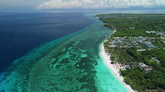 Aerial pulls back and tilts up over Dumaluan Beach, showing reef flats, white sand, boats, resorts, and Bohol coastline