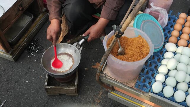 A street food vendor mixes cooked glutinous rice in a hot metal pan with seasoned dried coconut flakes. The aromatic coconut mixture is added to enhance the flavor and texture of the rice.