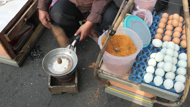 A street food vendor heats soaked glutinous rice in a small metal pan covered with a glass lid over a charcoal stove and fans charcoal to increase airflow.