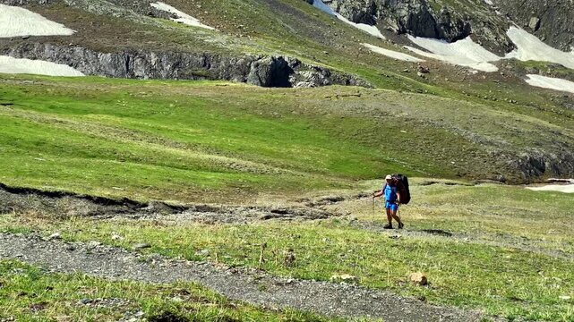 A solo hiker crosses an open alpine basin from Juta toward Roshka, carrying a loaded backpack through green meadows and patchy snowfields in Georgia, showing endurance, navigation, and trekking.