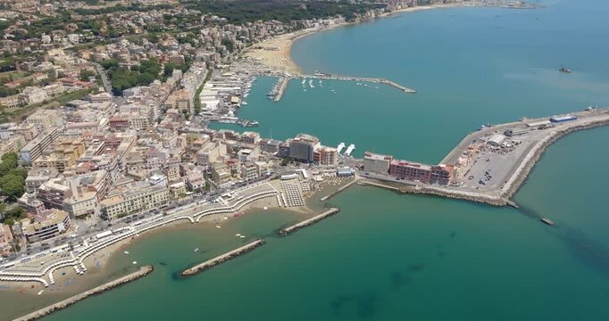 Panorama of Anzio, a city in province of Rome, Lazio, Italy. Aerial view of a sunny coastal town featuring a bustling harbor, long pier, turquoise sea waters, sandy beaches with umbrellas. 