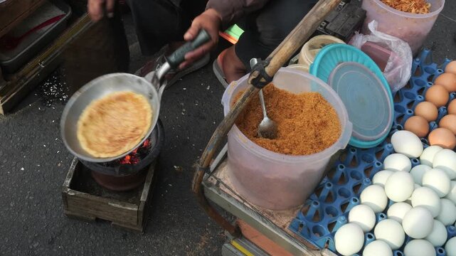 A street food vendor holds the pan upside down toward glowing charcoal while cooking Kerak Telor. This distinctive cooking method allows direct heat to reach the surface of the egg and rice mixture.