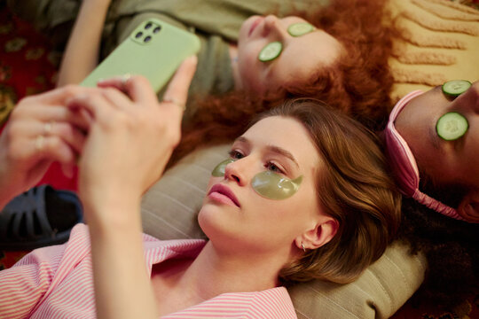 Young women relaxing with eye masks and cucumber slices, using smartphone during home spa session, expressing self care and friendship. Ideal for wellness, beauty, lifestyle marketing