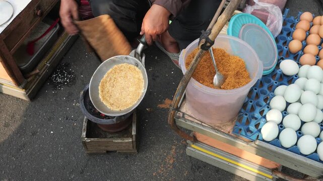 A street food vendor holds the pan upside down toward glowing charcoal while cooking Kerak Telor. This distinctive cooking method allows direct heat to reach the surface of the egg and rice mixture