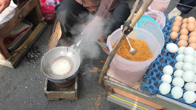 A street food vendor mixes partially cooked glutinous rice in a hot metal pan with seasoned dried coconut flakes. Adding Eggs, Dried Shrimp and Spices During Kerak Telor Preparation.