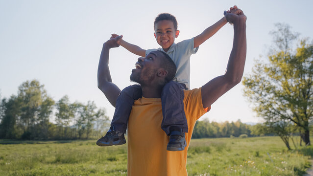 Father carries his son on his shoulders while they laugh and play in a green field at sunset. Quality time spent together in nature.