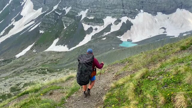 Hiker walking beside White Abudelauri Lake follows a narrow alpine trail beneath snow-covered Caucasus peaks, illustrating remote tourism, load carrying and trekking access in northern Georgia.