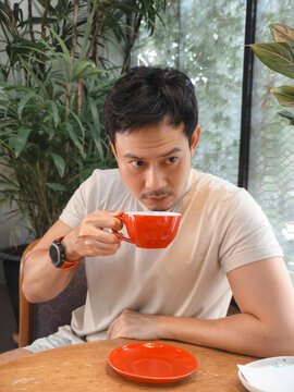 Thoughtful Asian man sips coffee from a vibrant red mug in a relaxed indoor setting.
