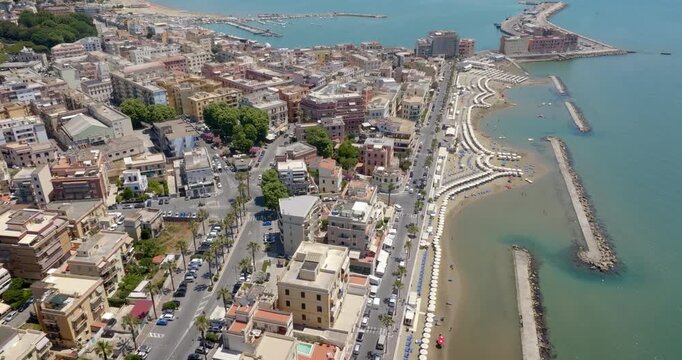 Aerial panoramic view of the coastal city of Anzio, in Lazio, Italy, showing its large harbor, long piers, sandy beaches with umbrellas, and urban buildings along the turquoise Mediterranean sea. 