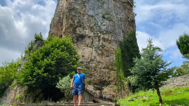 A traveler explores the landscaped grounds beneath Katskhi Pillar, where a secluded monastery crowns a towering limestone monolith, highlighting faith, heritage, geology, and rural tourism in Georgia