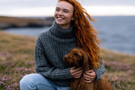 Close up portrait of redhead woman hugging her dog on coastal meadow for wallpaper and poster use highlighting warm smile and cozy knit sweater
