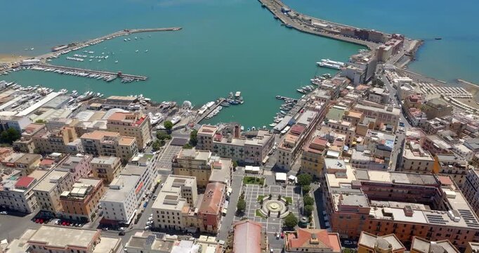 Aerial view of the harbor and coastal town of Anzio, located near Rome in Italy, featuring clear turquoise waters, breakwaters, docks with boats, and densely clustered buildings in a sunny summer day.