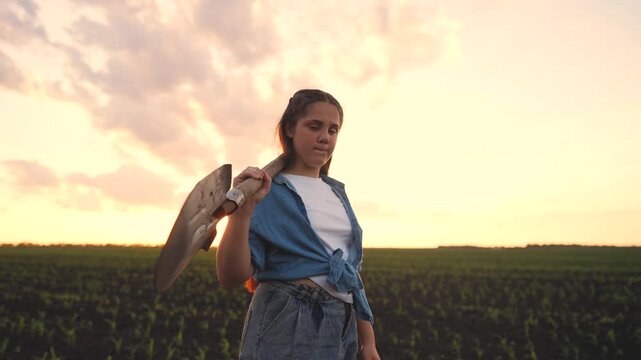 A teenage farmer girl stands in a field, holding a shovel as she gazes thoughtfully at the horizon. The teenage farmer girl appears determined amidst rows of young crops.
