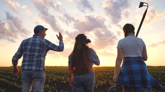 The man, girl, and woman walk together in the field, overseeing the sunset. The man gestures while the girl holds a shovel, and the woman carries a rake.