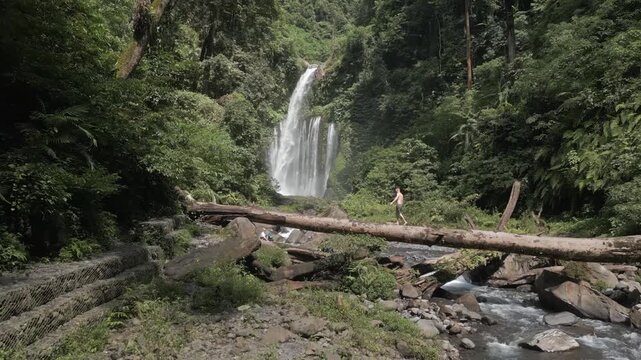 Man crossing a large fallen tree trunk over a flowing river in front of a majestic waterfall in tropical forest.
