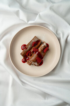 A plate holds a dessert featuring layers of chocolate sauce and fresh raspberries. The setting is simple, with a white tablecloth surrounding the dish