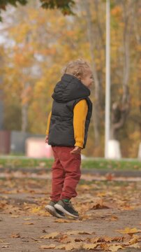 person walking and turning in outdoor area with fallen leaves and trees in the background, casual autumn scene with black vest over yellow shirt and red pants, side view.
