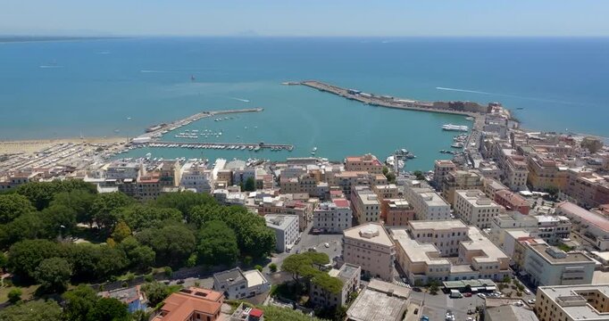 Aerial panoramic view of the port of Anzio, in Italy, with turquoise sea water, surrounding piers, docked boats, and urban coastal buildings extending toward the horizon under a sunny summer sky.