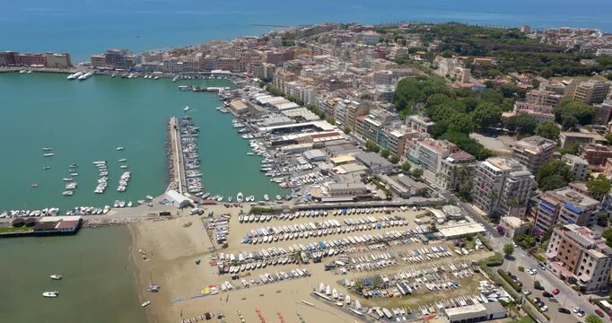 Aerial wide shot of the Anzio harbor and coastline, featuring vibrant sea waters, docked boats, sandy beaches, breakwaters, and urban coastal buildings in a summer day. It's a coastal town in Italy.