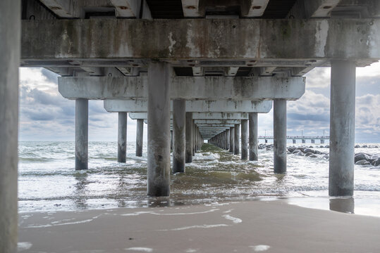 Symmetrical view beneath a seaside pier with concrete columns, gentle waves and dramatic cloudy sky. Coastal architecture and Baltic Sea landscape with strong leading lines.