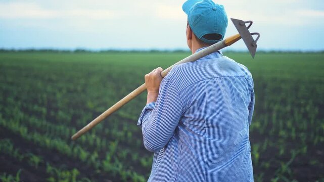A man stands in a field, holding a hoe with both hands. The man gazes out over the green landscape of crops and rich soil in the distance.