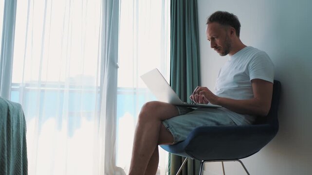 Remote professional types on laptop beside bright hotel window, representing bleisure travel, distributed teamwork, freelance mobility, and location-independent business.