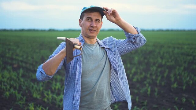 The man holds a hoe while standing in the field. The man stands amidst green crops under a cloudy sky, preparing for his work in the field.
