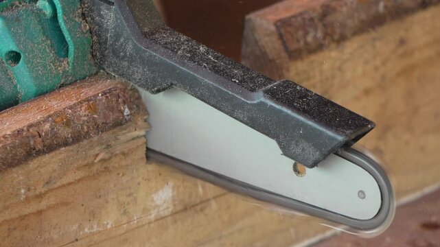 Electric chainsaw blade slices through pine board in a workshop, removing a rectangular notch from the wood. Extreme close up static shot.