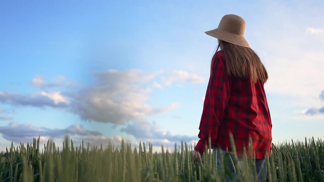 A woman in a red plaid shirt walks through a green field while wearing a straw hat. The woman enjoys the scenic view under a blue sky filled with clouds.