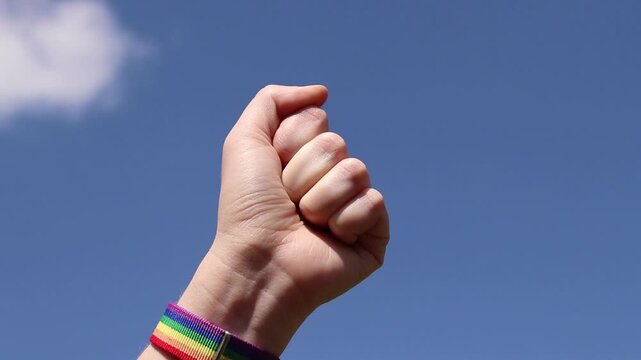 Raised fist with rainbow bracelet against blue sky with clouds. Symbol of fight for equal rights. Female hand with LGBTQ+ bracelet clenched into fist and raised to the sky