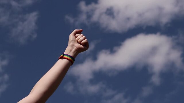 Raised fist with rainbow bracelet against blue sky with clouds. Symbol of fight for equal rights. Female hand with LGBTQ+ bracelet clenched into fist and raised to the sky