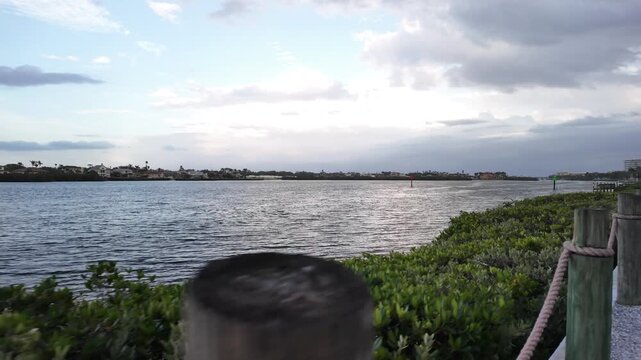 Smooth cinematic walking shot along a waterfront pathway beside a calm coastal lagoon with luxury condominiums, lush tropical landscaping, dramatic clouds, and peaceful evening light.