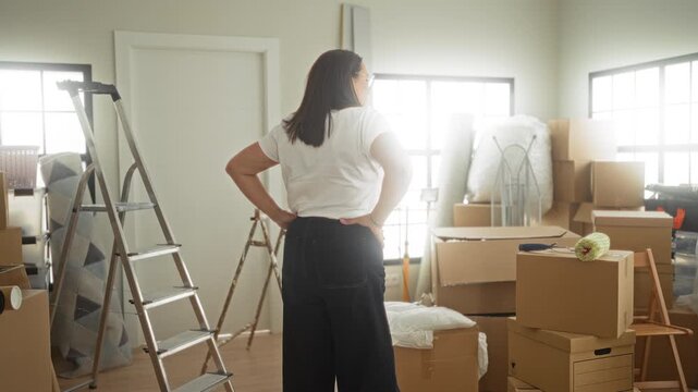 Woman hands on hips amid cardboard boxes, ladder, paint roller and wrapped furniture inside a building while surveying packed room; determination relocation.