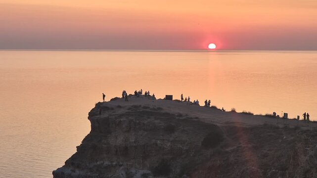 Sunset ocean cliff, people watch the golden sun dip below the horizon over calm sea, a beautiful scenic view