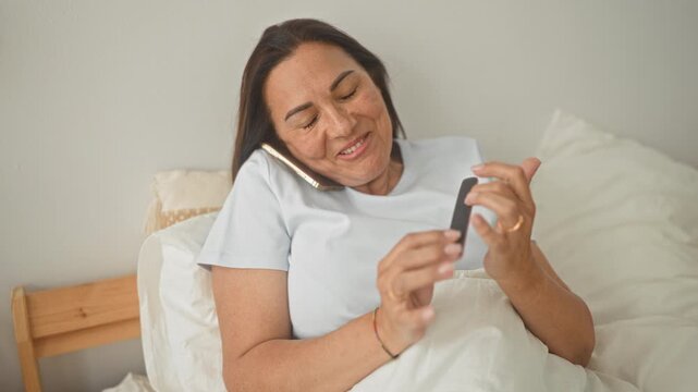Woman filing nails with emery board while holding smartphone to ear on bedroom bed; relaxed self care.