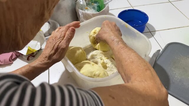 An elderly Latina woman is kneading dough to prepare corn arepas for breakfast in a bowl inside her kitchen in Neiva, Huila, Colombia. Concept of Colombian food and handmade