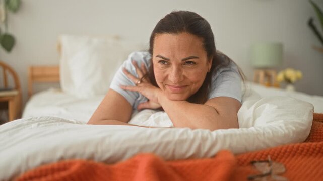 Middle age hispanic woman lying on bed propped on forearm, hand under chin smiling, orange throw and glasses visible in bedroom; serenity comfort warmth.