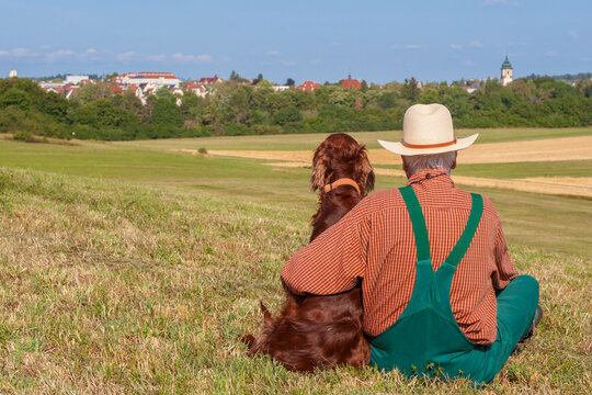 An older man in green dungarees sits on a grassy hill, sharing an affectionate moment with his loyal Irish Setter dog while looking out over a peaceful countryside village in the warm July sun.