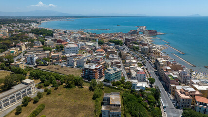 Fototapeta premium Aerial panoramic view of the sunny promenade and coastline in Anzio, in province of Rome, Italy. There are beachfront buildings, sandy shores, breakwaters, and clear sea under a bright summer sky.