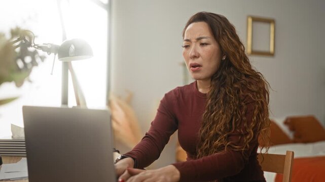 Woman covering mouth with hands while leaning over laptop at a desk in a building with bed and lamp visible; anxiety.