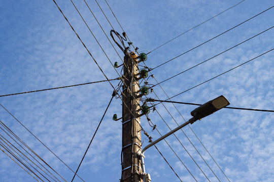 Wooden utility pole with multiple power lines and green insulators against a blue sky with scattered clouds, streetlight visible on the side