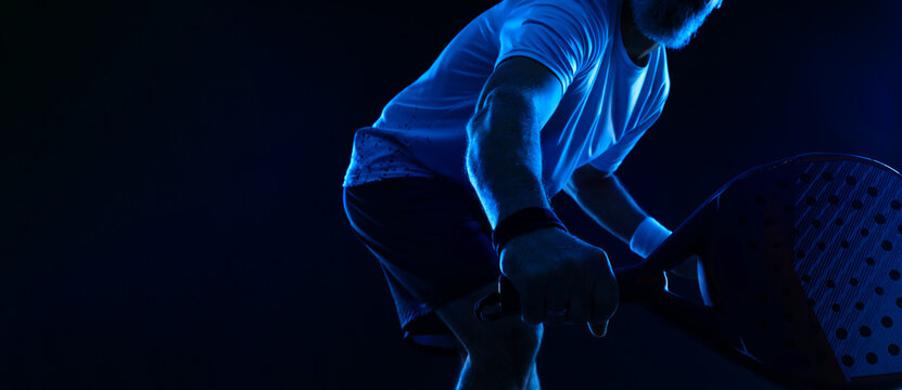 Close up Padel tennis player with racket. Man athlete with paddle racket on court with neon colors. Sport concept. Download a high quality photo for the design of a sports app or betting site.