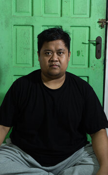 An adult Asian male sits calmly on a white mattress, looking directly into the camera lens with a very serious and unflinching facial expression. The close up lifestyle portrait features