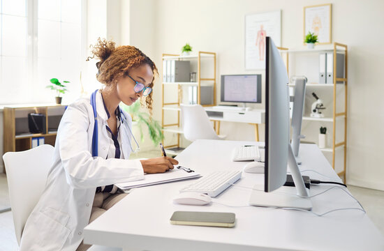 Doctor female physician computer clinic workstation. She writes notes and updates electronic records with analysis. Bright healthcare office with monitor. Clear documentation and consultation.